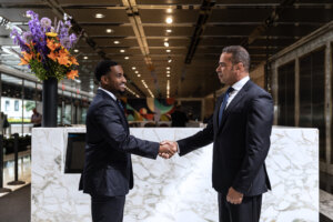 Two men in suits shake hands in a modern office reception area.