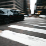 A black SUV moving quickly through a blurred urban intersection, surrounded by tall buildings, suggests motion and speed. The SUV is in sharp focus against the city's backdrop, while crosswalk lines appear prominently in the foreground—reminiscent of security companies ensuring swift mobility in urban landscapes.