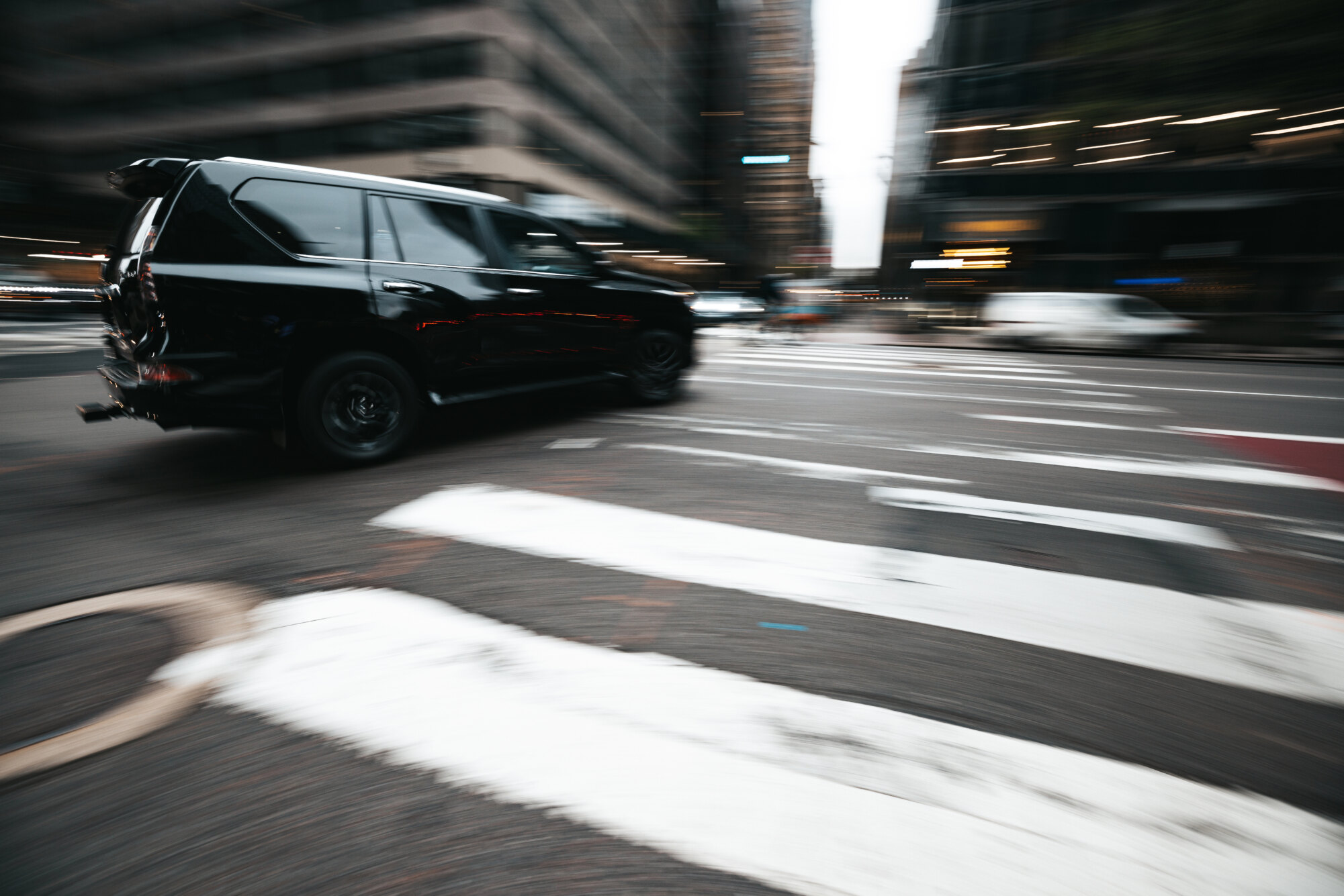 A black SUV moving quickly through a blurred urban intersection, surrounded by tall buildings, suggests motion and speed. The SUV is in sharp focus against the city's backdrop, while crosswalk lines appear prominently in the foreground—reminiscent of security companies ensuring swift mobility in urban landscapes.