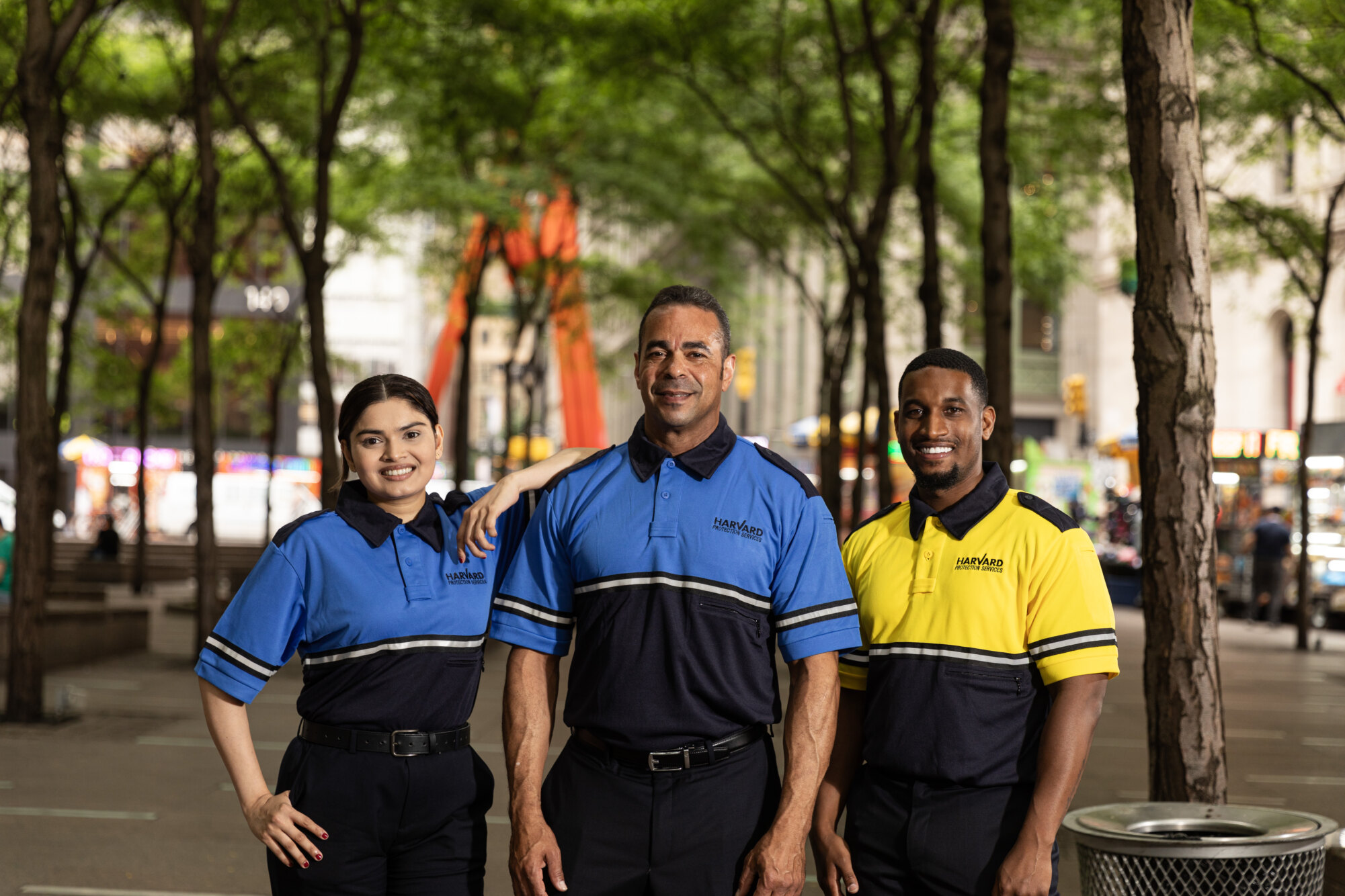 Three people in uniform pose outdoors on a tree-lined street with buildings in the background.