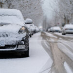 Snow-covered cars parked along a snowy, tree-lined street.