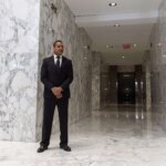 A person in a suit stands in a marble-floored hallway with elevators.