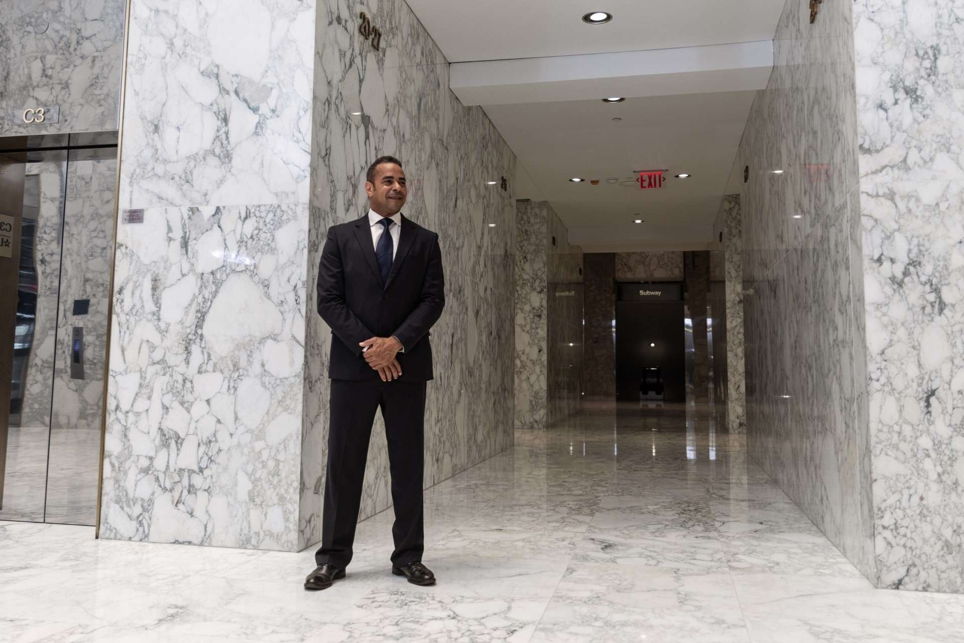 A person in a suit stands in a marble-floored hallway with elevators.